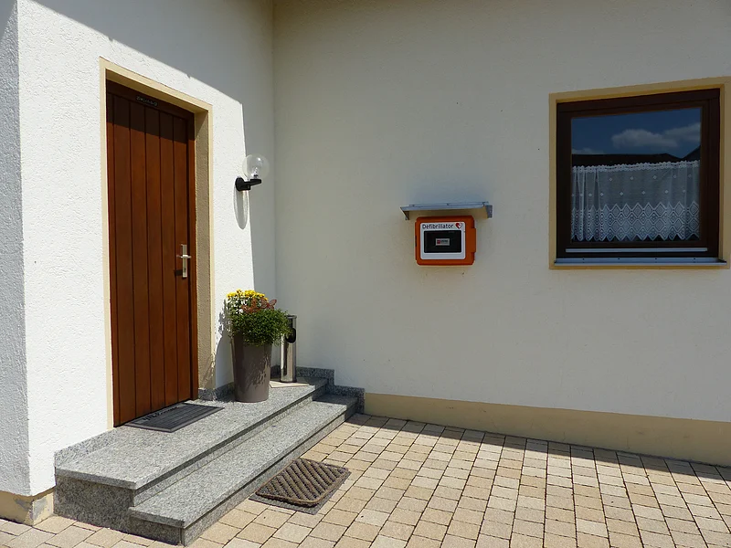 Eingangsbereich eines Hauses mit weißer Fassade, einer brauner Holztür, zwei Stufen davor, einem bepflanzten Blumentopf, einem Fenster mit Gardinen rechts im Bild und einem Defibrillator in einem orangen Kasten, der an der weißer Wand angebracht ist.