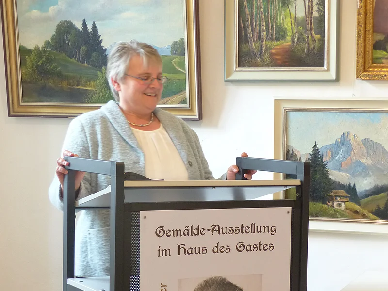 2. Bürgermeisterin grüßt Gäste und würdigt das Engagement Frau steht an einem Rednerpult mit einem am Pult angebrachtem Plakat zur Gemälde-Ausstellung von Hans Mayer im Haus des Gastes. Hinter Ihr an der weißen Wand sind Gemälden von Landschaften zu sehen.