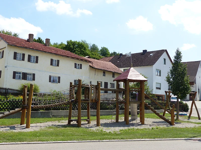 Spielplatz mit einem großen Klettergerüst aus Holz und einer Nestschaukel aus Holz auf grüner Wiese mit Kieselsteinen und einem Baum, vor Wohnhäusern und blauem Himmel am Tag.