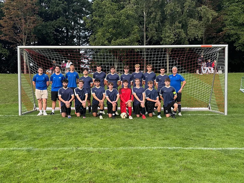 Fußballmannschaft mit 18 Spielern und 3 Trainern vor einem Tor auf Rasenplatz im Freien, Bäume im Hintergrund.