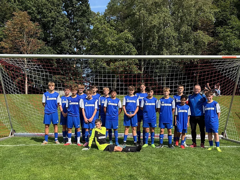 Jugendfußballmannschaft in blauen Trikots vor Tor auf grünem Spielfeld, im Hintergrund Bäume und zwei sitzende Personen.