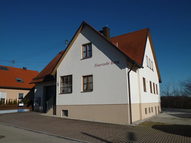 Feuerwehrhaus Itzing Weißes Feuerwehrgebäude mit rotem Dach, Aufschrift "Feuerwehr Izing" an der Fassade, gepflasterter Vorplatz, blauer Himmel. Links davon ein weiteres Haus in orange/beige mit rotem Dach. Vorderhalb gepflasterter Platz und blauer Himmel im Hintergrund.