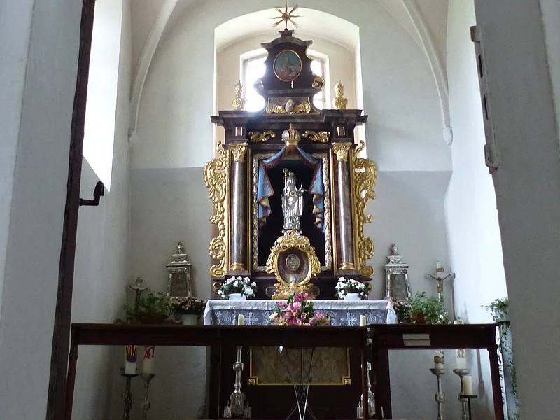 Walburgakapelle in der Stadtpfarrkirche St. Walburga Monheim Ein Blick durch eine Tür zu einem Kirchenaltar in der Walburgakapelle in Monheim, mit goldenen Verzierungen und einer Statue, der heiligen Walburga. Der Kirchenaltar ist umgeben von Kerzen und Blumen, in einem gewölbten Raum mit einer verzierten Decke.