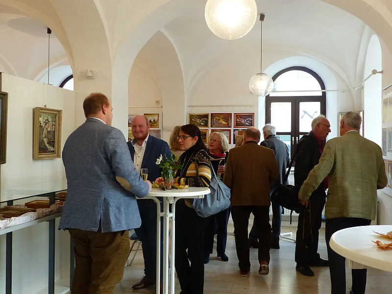 Blick in den Ausstellungsraum - zum 120. Geburtstag von Kunstmaler Hans Mayer Menschen in einem Ausstellungsraum mit weißen Wänden und Deckengewölben, die an Stehtischen sprechen und Kunstwerke betrachten.