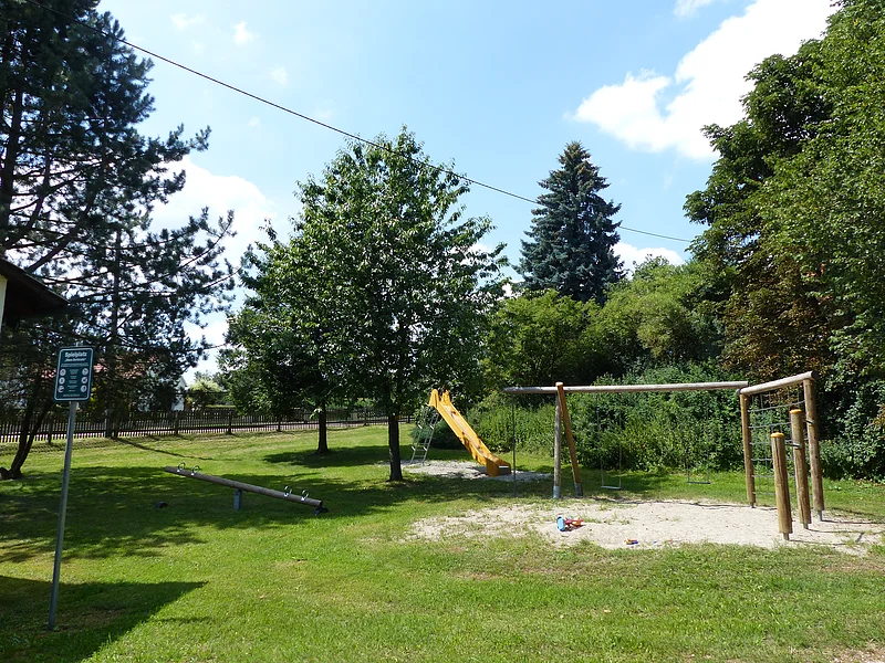 Großer Spielplatz mit einer Seilbahn, Schaukel, Rutsche, Wippe und Sandfläche, umgeben von Bäumen und Wiese bei Sonnenschein. Links im Bild steht ein Pfosten mit einem grünen Schild, auf dem die Spielregeln draufstehen.