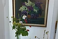Ölgemälde von Hans Mayer Stillleben von einem Fliederstrauß in einer Vase, das eingerahmt an einer weißen Wand hängt. Auf einem weißen Stehtisch im Vordergrund liegen Kastanien, Blätter und Zweige, Feldblumen in einer durchsichtigen kleinen Vase.