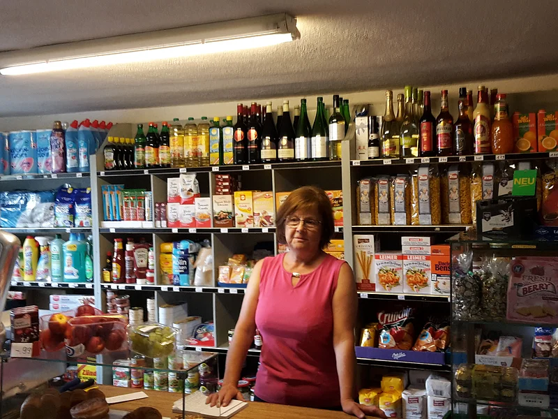 Frau in pinkem Oberteil steht hinter einer Ladentheke in kleinem Laden mit Regalen voller Lebensmittel und einer Waage an der Verkaufstheke links im Bild.