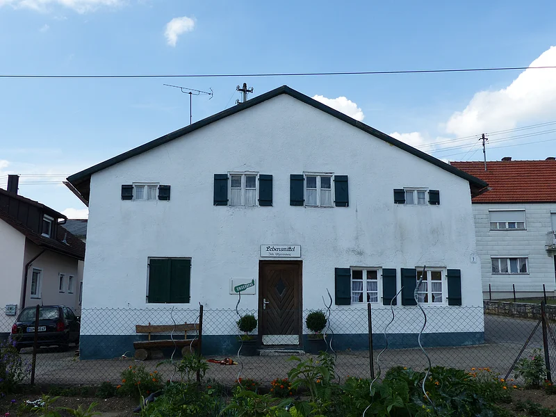 Weißes Haus mit grünen Fensterläden, einer braunen Holztür in der Mitte und einem weißen Schild darüber mit dem Schriftzug „Lebensmittel“. Im Vordergrund ist ein bepflanzter Garten mit einem Drahtzaun. Im Hintergrund weitere Häuser links und rechts und blauer Himmel.