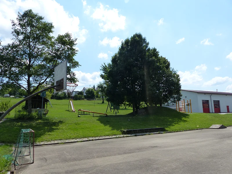 Spielplatz mit einem anliegenden Basketballkorb und einem kleiner Fußballtor darunter im Vordergrund. Im Hintergrund Spielplatz mit Rasenfläche mit Spielplatzgeräten wie Rutsche, Wippe, Klettergerüst, Schaukel und einem Trampolin umgeben von Bäumen bei sonnigem Wetter. Rechts daneben im Bild ein weißes Gebäude mit roten Toren.
