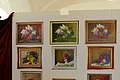 Blumengemälde vom Monheimer Kunstmaler Hans Mayer Neun gerahmte Gemälde mit Blumen und Früchten an weißer Wand, orange Blume im Vordergrund.