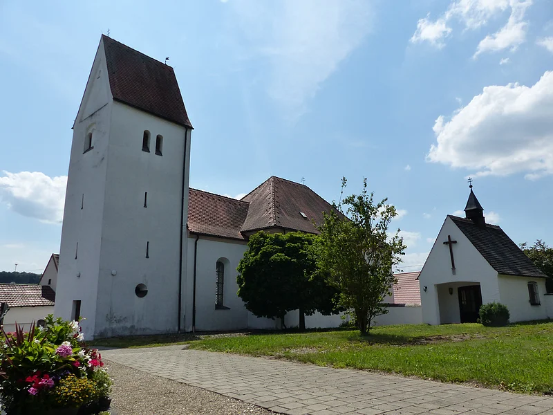 Filialkirche mit rotem Dach und einer kleiner Kapelle daneben mit einem Turm bei blauem Himmel und grünem Rasen im Vordergrund. Neben dem Rasen verläuft ein gepflasterter Gehweg mit Schottersteinen daneben.