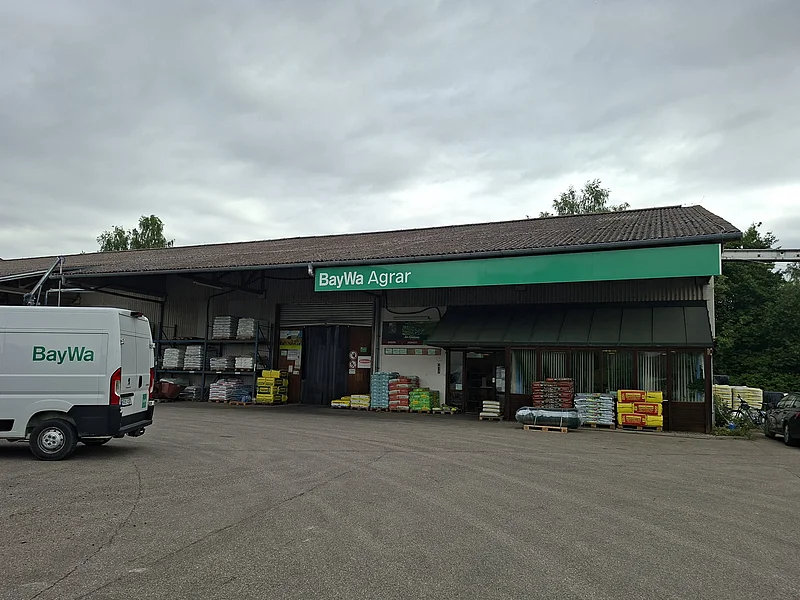 Außenansicht eines BayWa Agrar Gebäudes mit geparktem weißen Lieferwagen und Paletten mit Agrarprodukten davor.