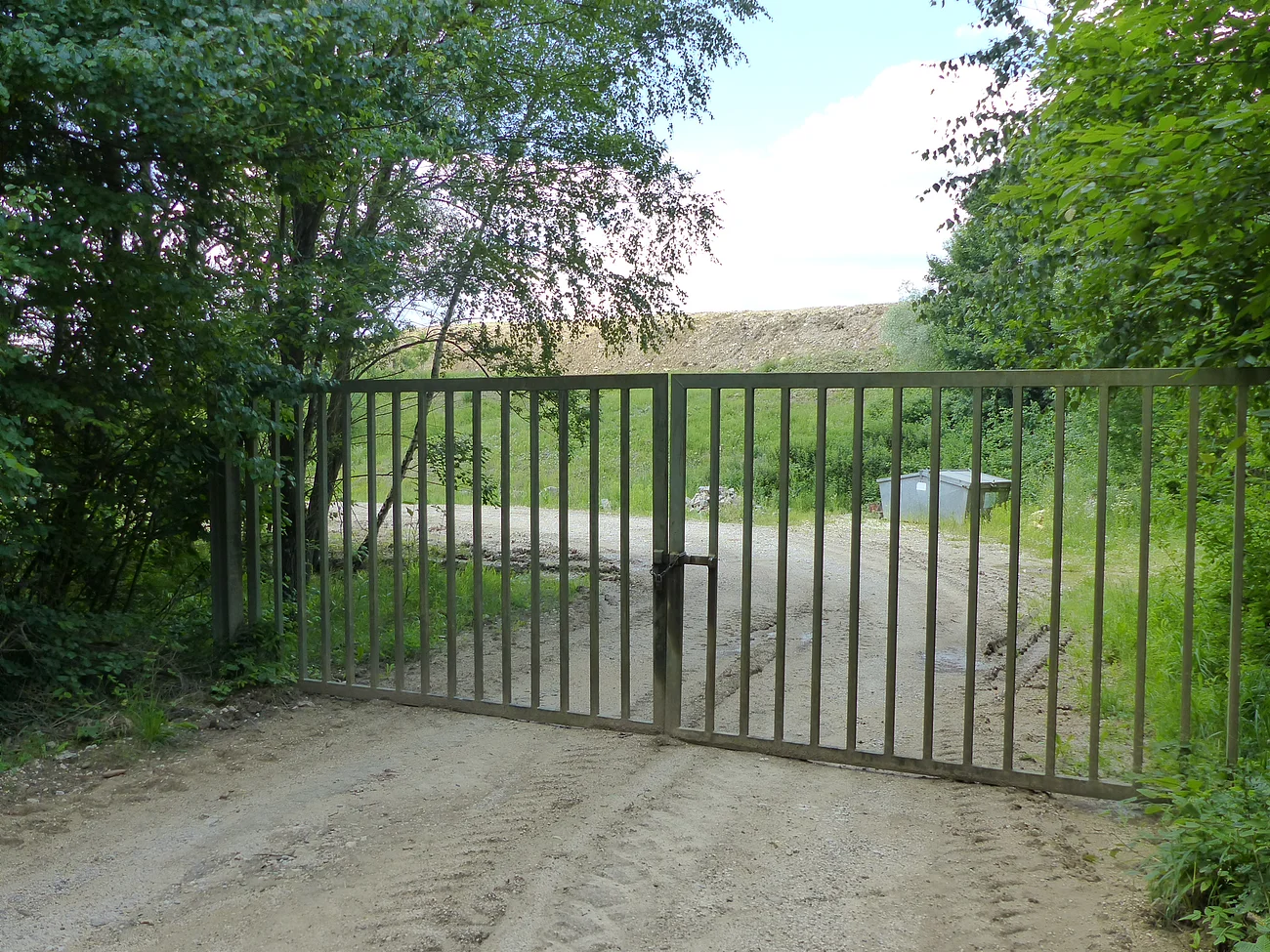 Erdaushubdeponie Monheim Verschlossenes Metalltor auf einem unbefestigten Weg, umgeben von Bäumen und Gras. Im Hintergrund ist ein Müllcontainer und ein Hügel sichtbar.