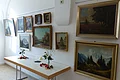 Im Mittelpunkt das gespendete Gemälde von Jos Bäuerle Gemälde an einer weißen Wand, an denen mehrere gerahmte Landschaftsbilder hängen, darunter ein Tisch mit Blumen.