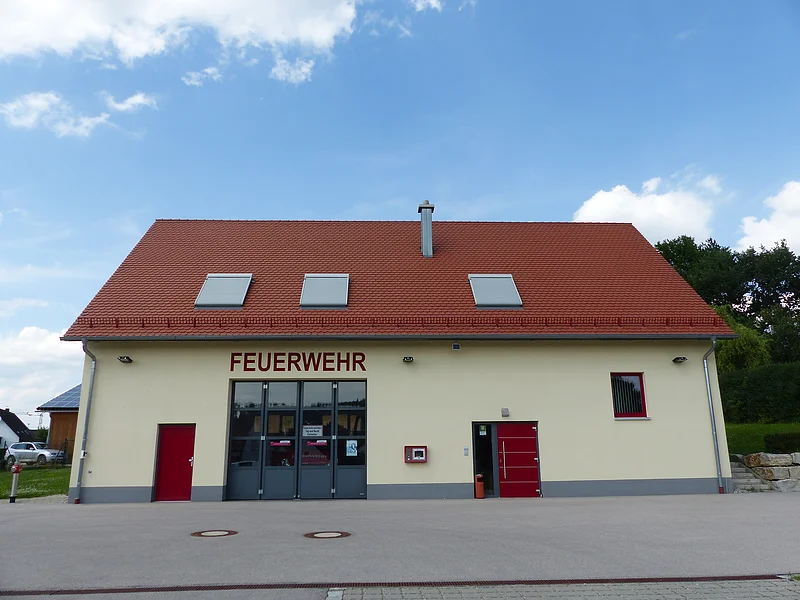 Gelbes Feuerwehrhaus mit rotem Satteldach, drei Dachfenstern und zwei Eingängen und einem Tor. Die Eingangstüren sind beide rot. Um das Gebäude große asphaltierte Fläche. Im Hintergrund blauer Himmel mit Bäumen und einem parkendem Auto.