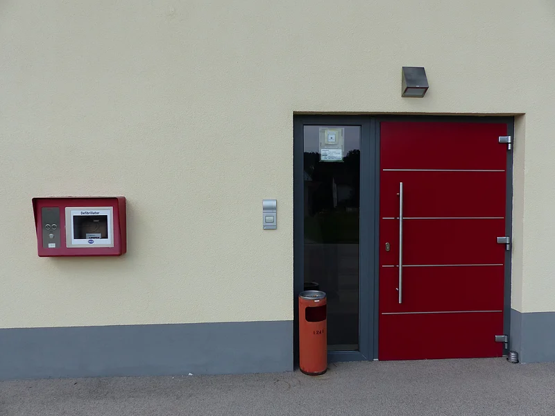 Gelbe Fassade eines Hauses mit roter Eingangstür mit Glasfenster. Links davon hängt ein roter Defibrillator-Kasten an der Wand und fein hoher roter Mülleimer steht vor der Haustür.