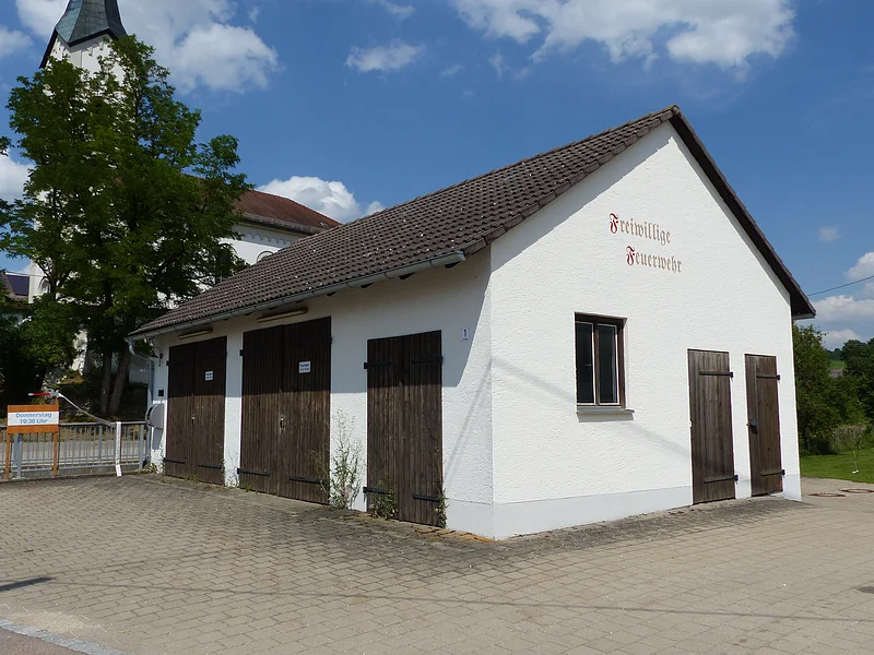 Weißes Gebäude mit braunem Dach und braunen Holztüren, darauf ein Schriftzug „Freiwillige Feuerwehr“ in braun/rot auf einem gepflastertem Platz. Im Hintergrund ein hoher Baum und eine Kirche mit Turm unter blauem Himmel zu sehen.