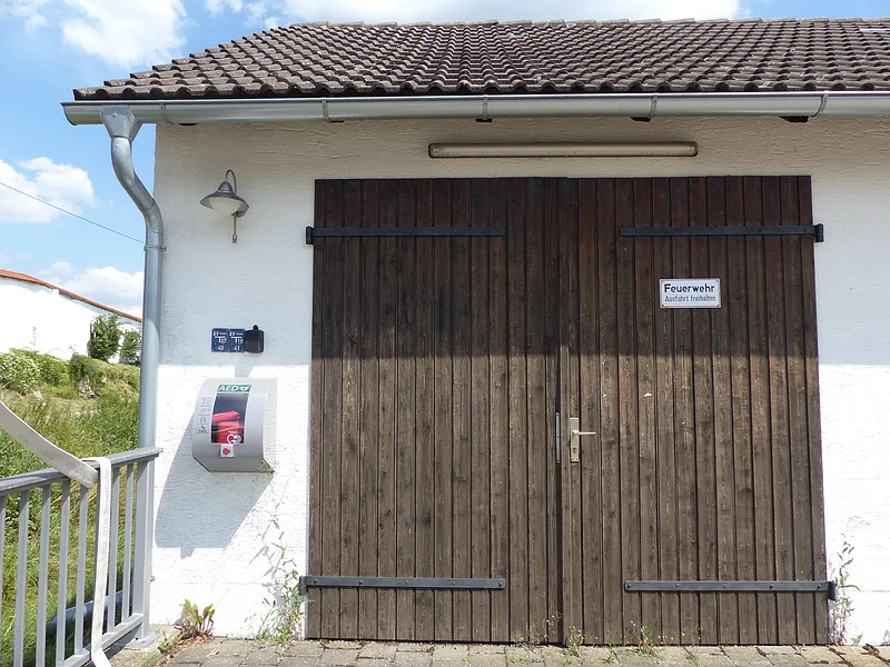 Braunes Holztor eines weißen Gebäudes mit weißem Schild drauf „Feuerwehr - Ausfahrt freihalten“ und automatischem externen Defibrillator an der Wand. Links im Bild im Hintergrund ein weißes Haus mit grüner Wiese bei blauem Himmel.