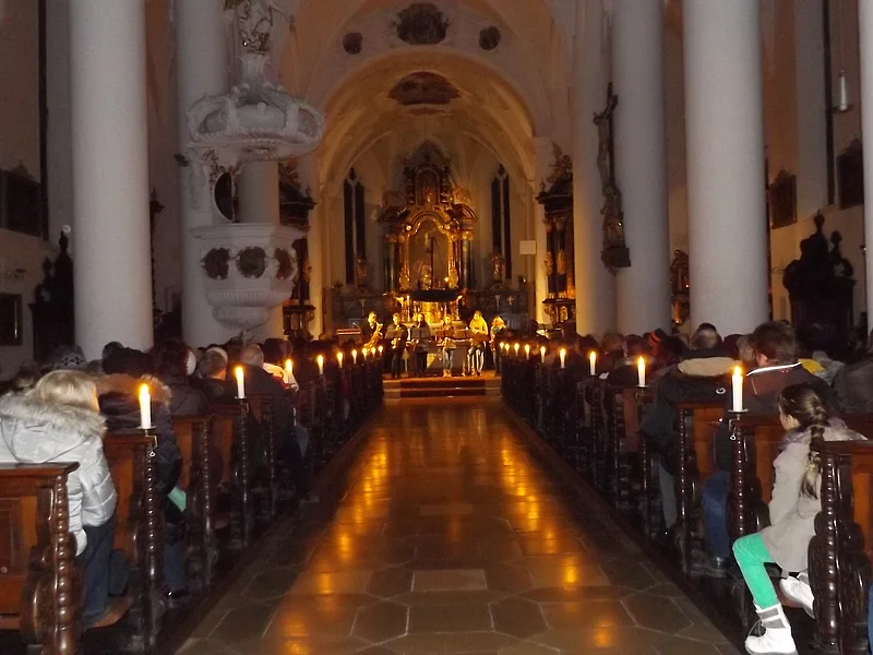 Innenraum einer Kirche mit Kerzen beleuchtet, Menschen sitzen auf Holzbänken, Blick auf den Altar im Hintergrund.