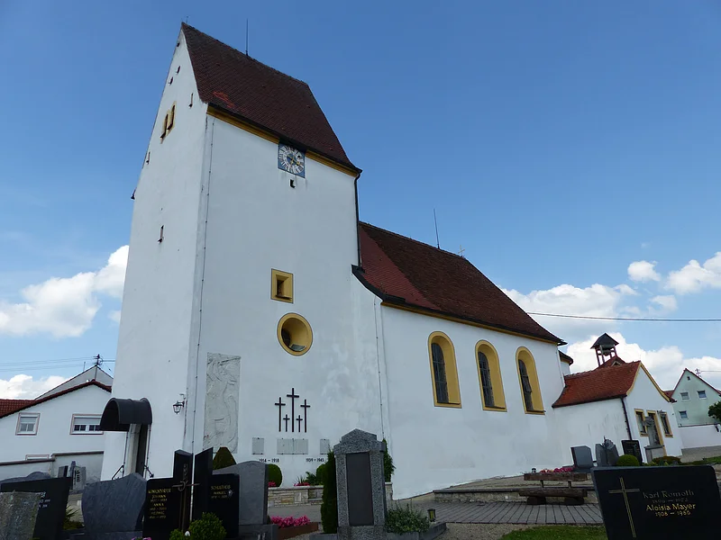 Weiße Pfarrkirche mit Turm, gelb eingerahmten Fenstern, einer Uhr und einem anliegenden Friedhof mit Grabsteinen, unter blauem Himmel.