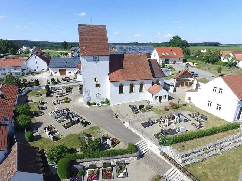 Luftaufnahme einer Kirche mit Friedhof und umliegenden Häusern bei klarem Himmel.
