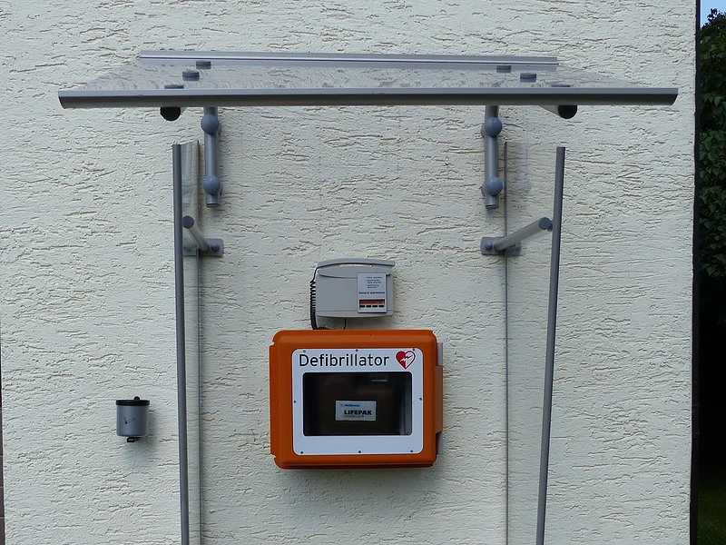 Defibrillator in orangefarbener Box an weißer Außenwand unter transparentem Wetterschutzdach befestigt.