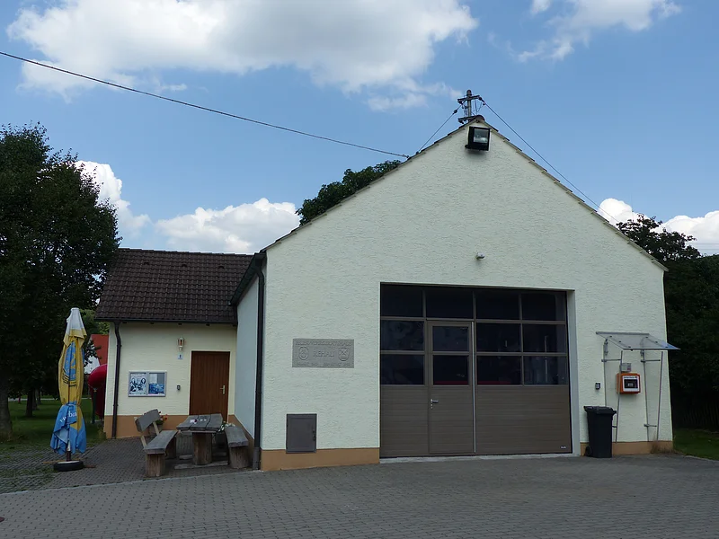 Gelbes Gebäude mit braunem Dach und großem braunem Tor, mit zwei Holzbänken und einem Tisch links vom Gebäude und einem geschlossenen Sonnenschirm, vor blauem Himmel mit Wolken. An der Hauswand rechts im Bild ein befestigter Defibrillator unter durchsichtigem Wetterschutzdach.