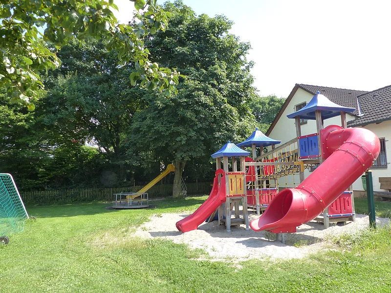 Spielplatz mit zusammenhängenden Spielgeräten wie zwei roten Rutschen, Klettergerüst und Sandfläche unten, neben einem weißen Wohnhaus. Im Hintergrund eine weitere gelbe Rutsche mit einem Karussell umgeben von Bäumen. Links im Bild teilweise ein grünes Fußballtor zu sehen.