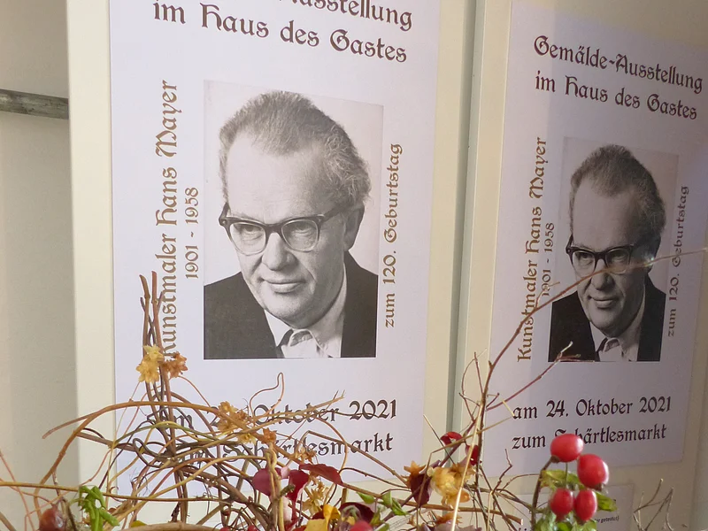 Gemäldeausstellung - Haus des Gastes 24.10.2021 Zwei Plakate mit je einem Porträt von Kunstmaler Hans Mayer darauf und der Ankündigung einer Gemälde-Ausstellung am 24.10.2021. Im Vordergrund liegen bunte Herbstblumen.