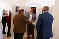 Ehrengäste bei der Eröffnung der Kunstausstellung Im Vordergrund stehen drei Männer an einem Stehtisch mit Sektgläsern vor Ausstellungstafeln mit Fotos und viel Text, in einem hellen Raum. Im Hintergrund zwei weitere Personen vor Gemälden an der Wand.
