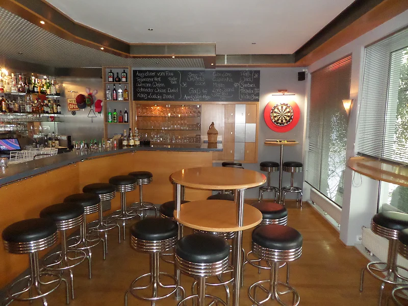 Innenansicht einer Bar mit Hockern, Holztischen, einer Dartscheibe und einer schwarzen Getränketafel. Links im Bild Regale mit vielen Flaschen und Gläsern. Rechts zwei Fenster mit geschlossenen Jalousien.