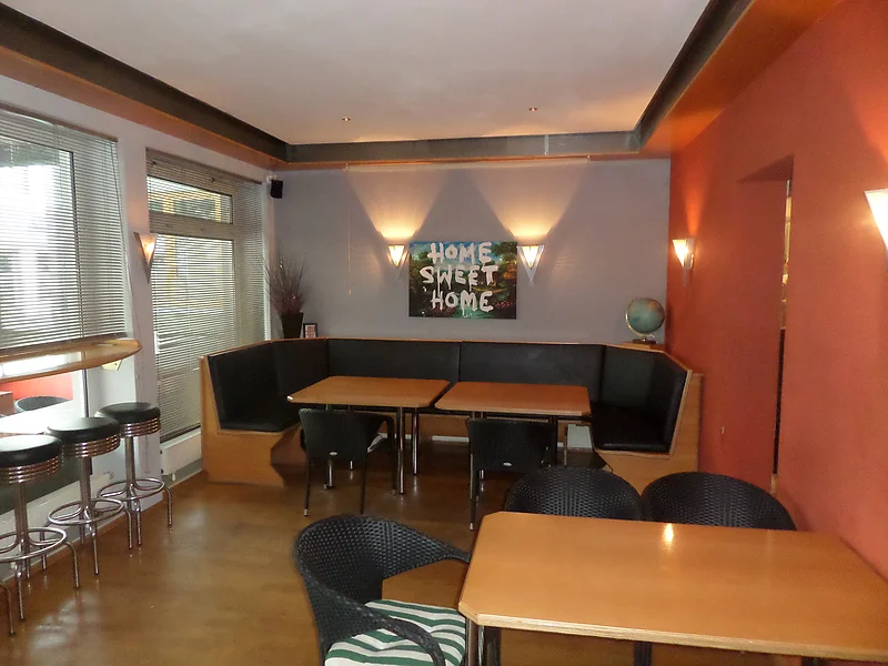 Innenraum einer Bar mit Holztischen, schwarzen Stühlen, drei Barhockern und einer Eckbank. Fenster mit Jalousien und Wandbild mit Schriftzug „Home Sweet Home“ auf einer grauen Wand. Rechts eine orangefarbene Wand mit angebrachten Leuchten.