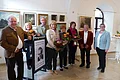 Gemälde-Ausstellung zum 120. Geburtstag des Monheimer Kunstmalers Hans Mayer Acht Personen stehen in einem Raum nebeneinander mit Gemälden an den weißen Wänden, vier Frauen davon halten Blumensträuße, davor ein Rednerpult mit einem Schild mit einem Porträt darauf.