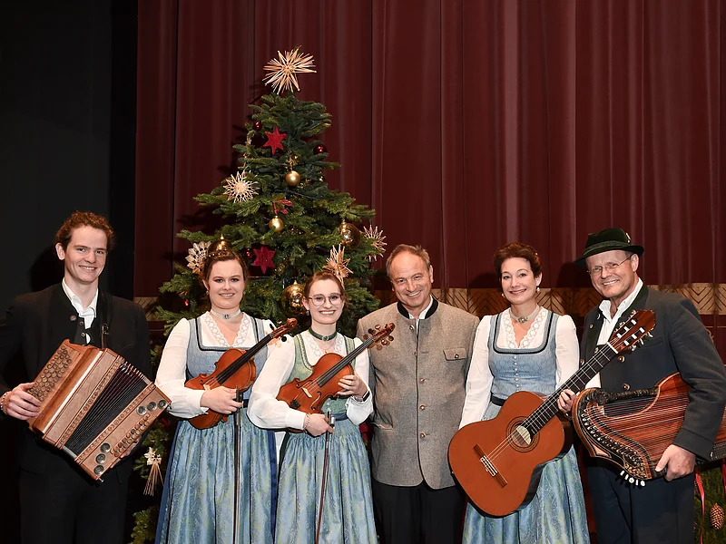 Pfarrer R.M. Schießler Hl. Nacht -Familienmusik Rehm & Rupert Biegel Sechs Personen in Trachten vor geschmücktem Weihnachtsbaum mit Musikinstrumenten posieren vor rotem Vorhang. Heilige Nacht mit Pfarrer R.M. Schießler & Familienmusik Rehm & Rupert Biegel am 21.12.2026 in der Monheimer Stadthalle