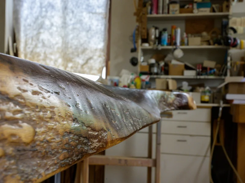 Möbel Meßmer - Tisch seitlich Nahaufnahme einer unregelmäßig geformten dunklen Holzoberfläche eines Tisches in einer Werkstatt mit Regalen und Werkzeugen im Hintergrund.