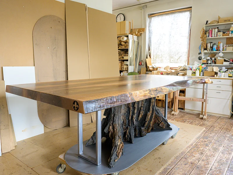 Möbel Meßmer - Tisch Dunkler Holztisch mit Baumstamm als Tischbein in Werkstatt mit Werkzeugen und Holzplatten im Hintergrund.