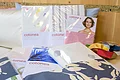 Möbel Meßmer - Textilien Mehrere verpackte Textilien der Marke "Cotonea" auf einem weißen Sofa, daneben eine Schale mit bunten Stoffmustern, im Hintergrund eine Holzwand.