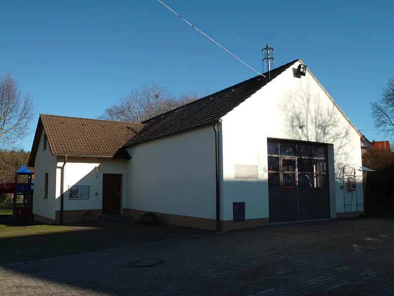 Ein einstöckiges Gebäude mit weißer Fassade und braunem Dach, das Feuerwehrhaus in Rehau steht auf einem gepflastertem Platz. Links eine Tür, rechts ein großes Tor. Im Hintergrund blauer Himmel und Bäume.