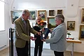 Familie Bäuerle schenkt Gemälde des Kunstmalers Hans Mayer Drei Personen nebeneinander in einem Raum mit mehreren Gemälden, die im Hintergrund an den weißen Wänden hängen, eine Frau hält Blumen, eine andere übergibt eine Geschenkbox an einen Mann.