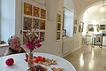 Blick in die Ausstellung zum 120. Geburtstag des Monheimer Kunstmalers Hans Mayer Älterer Mann sitzt hinter einem Stehtisch mit Blumen, Kastanien, Blättern und Apfel in einem hellen Raum mit Gemälden an den weißen Wänden. Im Hintergrund sind Rundbögen an der Decke zu sehen.