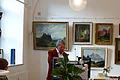Eröffnung der Kunstausstellung Älterer Mann im braunem Sakko und Brille liest an einem Rednerpult stehend vor, im Vordergrund ein Stehtisch mit Sektgläsern, Blumen und Herbstblättern. Im Hintergrund Gemälde an der weißen Wand und ein weiterer dekorierter Tisch.