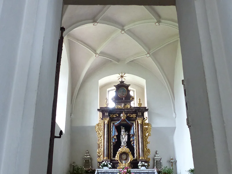 Ein Blick durch eine Tür zu einem Kirchenaltar in der Walburgakapelle in Monheim, mit goldenen Verzierungen und einer Statue, der heiligen Walburga. Der Kirchenaltar ist umgeben von Kerzen und Blumen, in einem gewölbten Raum mit einer verzierten Decke.