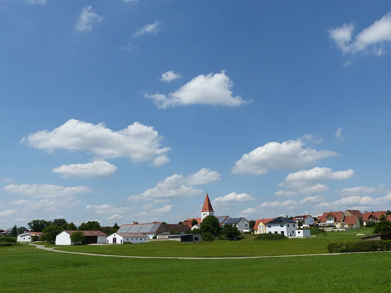 Flotzheim Dorf mit Kirche, Häusern und einer Feldstraße vor grünem Feld unter blauem Himmel mit weißen Wolken.