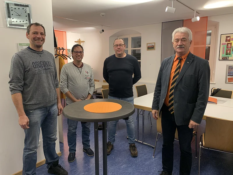 Vier Männer stehen nebeneinander in einem Raum um einen schwarzen Stehtisch mit oranger Unterlage drauf, einer von ihnen trägt einen Sakko, mit orangenem Hemd und einer Krawatte. Im Hintergrund weitere Tische und Stühle, schwarz-orange Vorhände an Fenstern, Bilder an Wänden und eine Uhr.