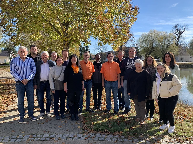Gruppenfoto von 16 Personen im Freien vor einem großen Baum und einem Teich rechts im Bild, an einem sonnigen Herbsttag.